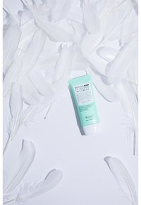 Benton UV Defense Solkräm i en turkos tub med texturerad yta, placerad bland vita fjädrar på en ljus bakgrund.