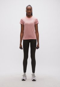 Γυναίκα σε όρθια στάση, κρατώντας βαράκια 1 κιλού σε κάθε χέρι, φορώντας ροζ μπλούζα Puma, μαύρο κολάν και λευκά αθλητικά παπούτσια σε απλό φόντο.