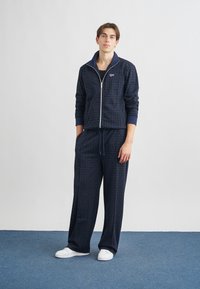 Jūras spēku zip-up treniņtērps ar ribotām aprocēm un jostasvietu, ar visā virsmā esošu logo rakstu. Saskaņots ar baltām sporta apaviem kontrastam.