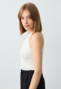 Jonge vrouw met schouderlang blond haar, gekleed in een witte mouwloze top en zwarte broek, die iets naar de camera kijkt tegen een witte achtergrond.