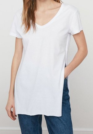 Vrouw draagt een losse witte V-hals top met korte mouwen en zijslitsen, gecombineerd met een blauwe spijkerbroek, staand tegen een effen lichte achtergrond.