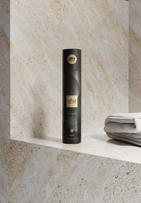 Boîte noire de shampoing sec ghd sur une étagère en pierre beige à côté de serviettes blanches pliées dans une salle de bain.