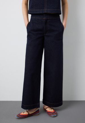 Person, die eine dunkelblaue, weit geschnittene Jeanshose trägt, mit den Händen in den Taschen und bunten, gehäkelten flachen Schuhen mit Knöchelriemen.