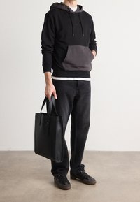 Sudadera negra con bolsillo frontal gris, jeans negros y zapatillas negras. Sosteniendo un elegante bolso de cuero negro con correas planas.