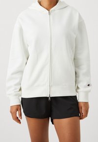 Sudadera blanca con cremallera hecha de tela suave, con capucha forrada, mangas largas y un pequeño logo en la manga izquierda. Combinada con pantalones cortos negros.