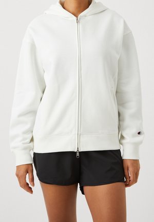 White zip-up hoodie made of soft fabric, featuring a lined hood, long sleeves, and a small logo on the left sleeve. Paired with black shorts.