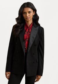 Melna divrindu žakete ar satīna klātbūtni, strukturētām plecu daļām un divām pogām. Valkāta virs sarkanas rūtainās blūzes ar kakla saišu.