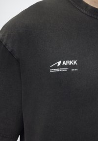 Μαύρο t-shirt με λευκό λογότυπο ARKK και κείμενο "Copenhagen Essentials ©Innovation Included EST 2015" στο στήθος.