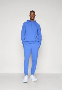 Blauwe katoenen hoodie met een voorzak en trekkoord, gecombineerd met bijpassende joggingbroek. Draagt witte sportschoenen.