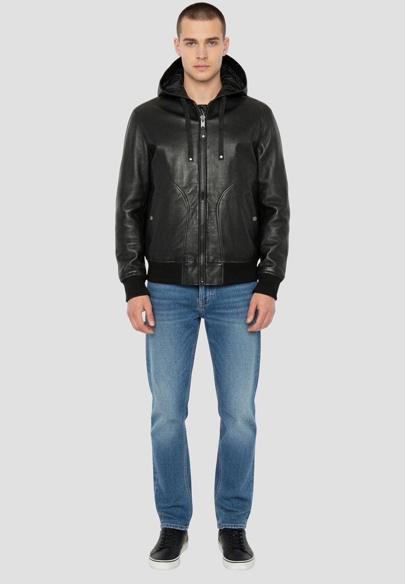 Veste en cuir noire avec capuche, fermeture éclair et poches avant. Portée avec un jean bleu clair et des baskets noires. Design classique décontracté.