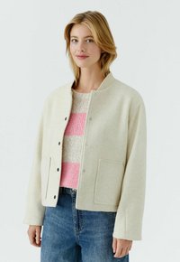 Frau trägt eine beige geknöpfte Jacke über einem rosa-cremefarben gestreiften Pullover mit blauen Jeans, steht vor einem weißen Hintergrund.