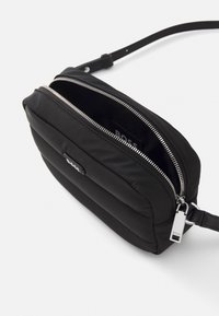 Černá nylonová crossbody taška s polstrovaným designem, stříbrnými zipy a kovovou značkovou visačkou. Disponuje jedním nastavitelným popruhem.