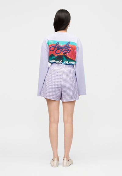 Γυναίκα με μακριά μαύρα μαλλιά που φορεί ένα μωβ πουκάμισο με μακριά μανίκια με πολύχρωμο σχέδιο βουνού και κείμενο στην πλάτη, μωβ σορτς με καρώ σχέδιο και μπεζ παπούτσια.