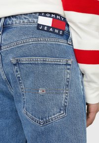 Modré džínové kalhoty s etiketou "TOMMY JEANS," červenými a bílými obdélníkovými akcenty, viditelným švem a dvěma zadními kapsami.