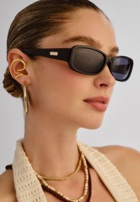 Γυναίκα με μαλλιά χτενισμένα προς τα πίσω, που φοράει μαύρα ορθογώνια γυαλιά ηλίου, μεγάλα χρυσά κρίκους σκουλαρίκια και στρω μένα κολιέ με χάντρες, με κρεμ μπλούζα.