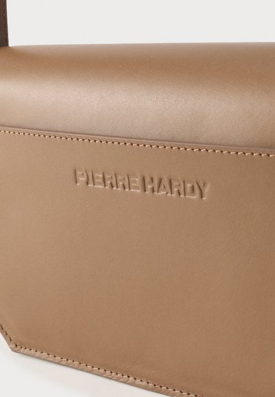 Κοντινό πλάνο τσάντας από ταμπά δέρμα με εμφανείς ραφές και το ανάγλυφο όνομα της μάρκας "PIERRE HARDY" στην επιφάνεια.