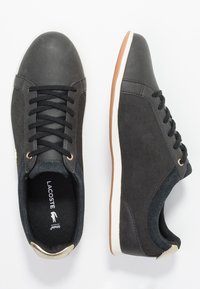Ett par svarta Lacoste-sneakers med vita sulor, svarta snören och bruna gummisålar visas från ovan och sidan mot en vit bakgrund.