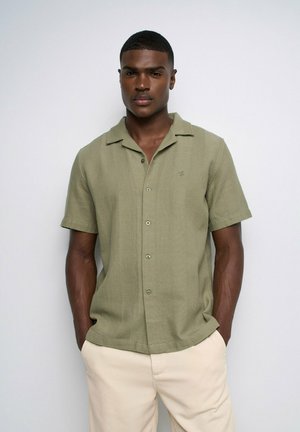 Hombre de pie con las manos en los bolsillos, vistiendo una camisa de manga corta color verde oliva con botones y pantalones beige claros sobre un fondo liso.