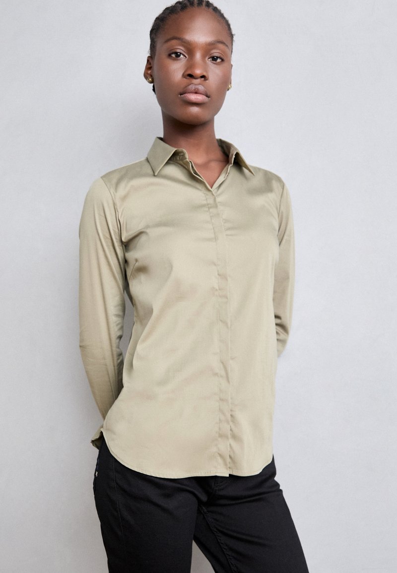 Khaki long-sleeve button-up shirt with a collar, smooth texture, and a curved hem, paired with black pants. Minimalistic design.