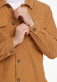 Katoenen overhemd in warm bruin, met verticale ribbels, zwarte knopen en een borstzak. De manchetten zijn met knopen dicht, wat een wit T-shirt eronder laat zien.