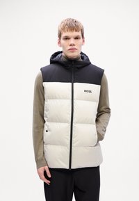 Polstret vest med svart hette, kremfarget front og merkevarelogo. Har glidelås og lommer, båret over en beige langermet skjorte.