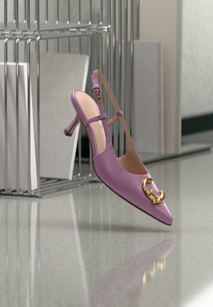 Paarse slingback hoge hak met gouden decoratieve gesp aan de voorkant, afgebeeld op een reflecterend oppervlak naast een metalen rek met papieren.