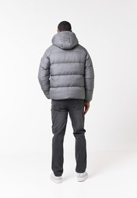 Chaqueta acolchada gris con capucha, con secciones acolchadas horizontales. Se lleva sobre jeans negros y zapatillas blancas. Diseño minimalista.