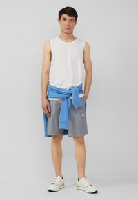 Witte mouwloze shirt, lichtblauwe sweatshirt om de taille geknoopt, grijze shorts met zakken en witte sneakers. Eenvoudige en casual look.