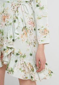 Abito a portafoglio floreale in verde pastello, con rouches lungo l'orlo e i bordi delle maniche. Realizzato in tessuto leggero, presenta fiori rosa e bianchi.