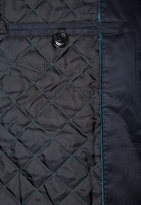 Svart quiltad jacka med insida i diamantmönster, kontrasterande tealfärgade kantband och svart knappdetalj. Slät syntetisk textur.