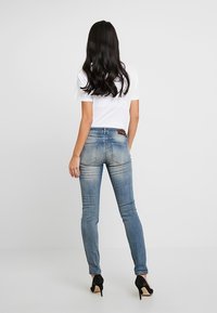 Vaqueros ajustados de color azul con un lavado desgastado, diseño de cinco bolsillos y un parche de cuero marrón en la cintura trasera. Se llevan con una camiseta blanca y tacones negros.