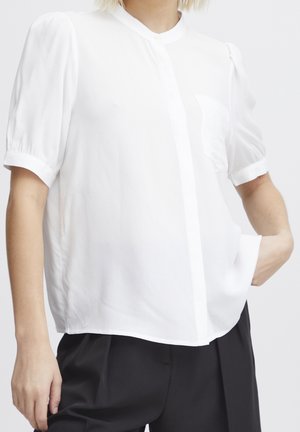 Kvinde iført en hvid kortærmet bluse med puffede skuldre og sorte bukser, poserer med den ene hånd i lommen mod en ensfarvet baggrund.