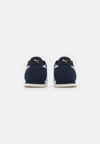 Zapatillas de ante azul marino con acentos blancos y un logo amarillo en el talón. Presentan una puntera redondeada y un diseño de perfil bajo.