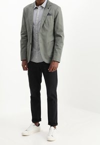 Mężczyzna w jasnoszarym żakiecie, szarej koszuli z guzikami, czarnych spodniach i białych sneakersach, stojący na jednolitym białym tle.