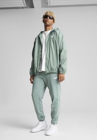 Lys grønn vindjakke med hvite detaljer, kombinert med matchende joggebukser. Hvite sneakers fullfører antrekket. Modellen har på seg solbriller.