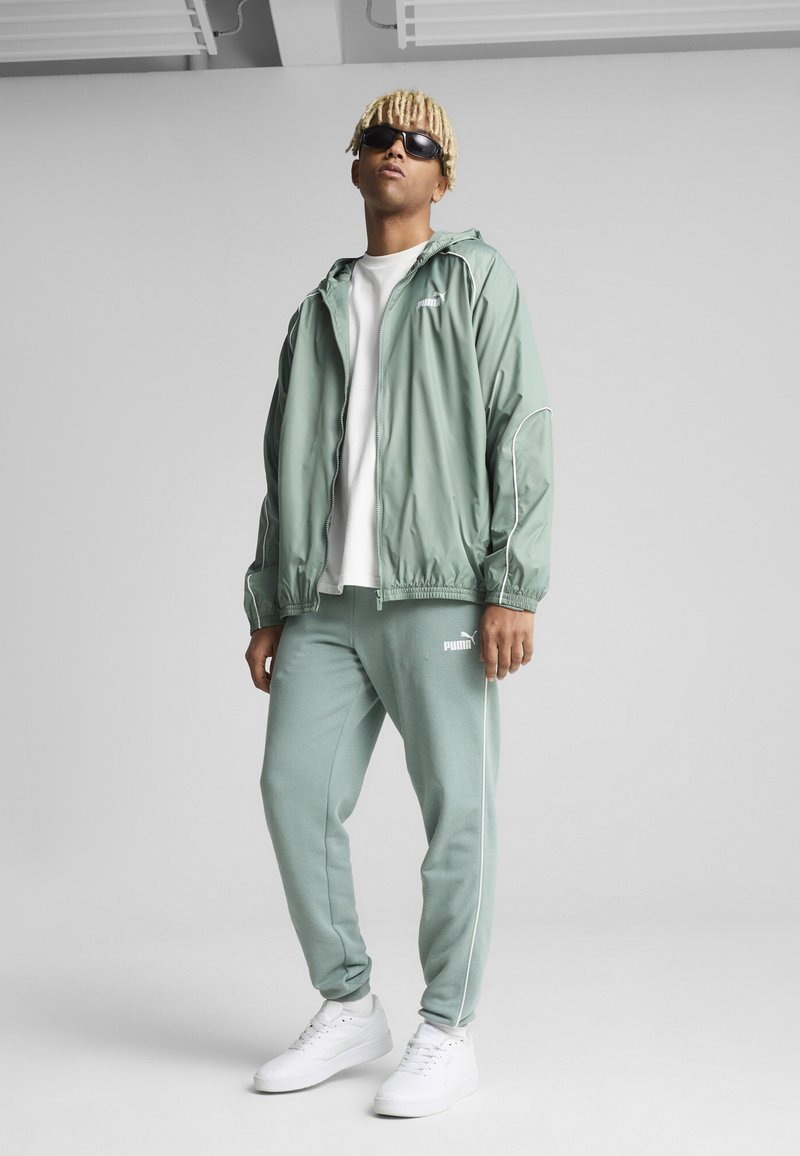 Hellgrüne Windjacke mit weißen Akzenten, kombiniert mit passenden Jogginghosen. Weiße Sneaker runden das Outfit ab. Das Modell trägt Sonnenbrillen.