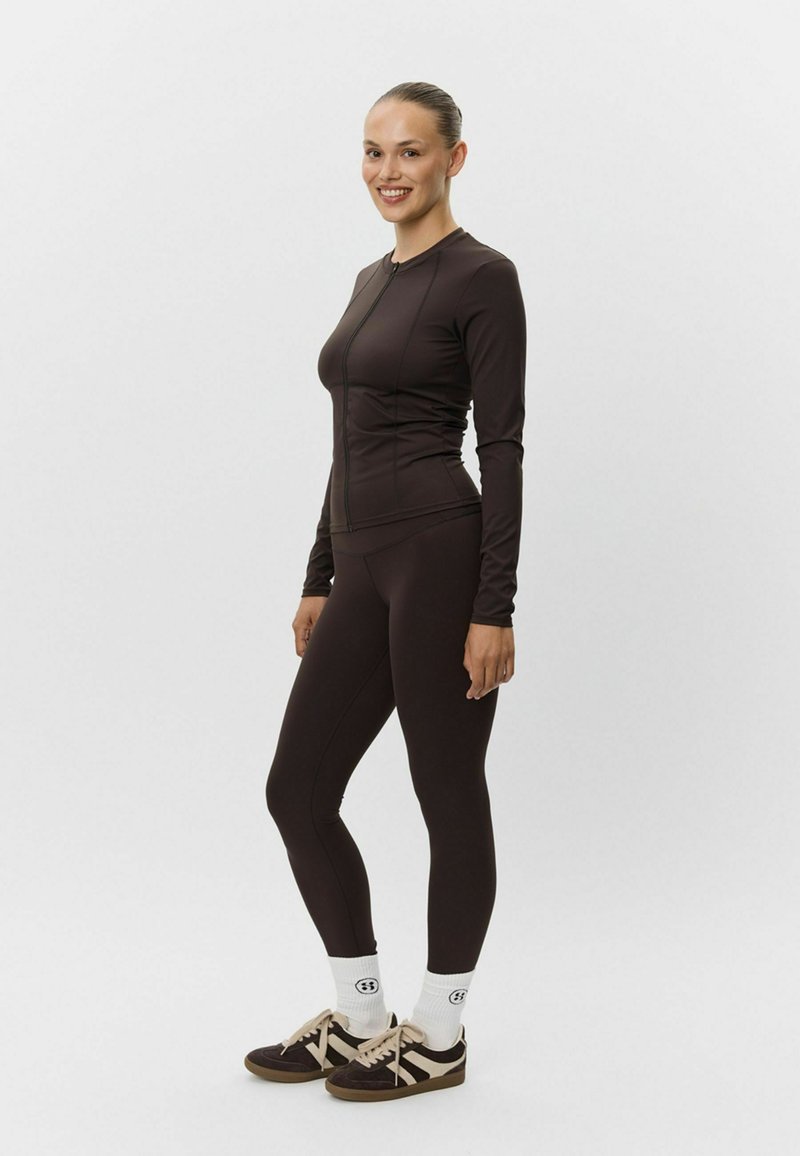 Tmavohnedý top s dlhým rukávom a zipsom vpredu, doplnený o zodpovedajúce legíny. Model má na sebe čierne tenisky s béžovými akcentmi a biele ponožky.