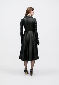 Μαύρο δερμάτινο φόρεμα με εφαρμοστή μπούστο και φούστα που φουσκώνει. Διαθέτει γιακά και μακριά μανίκια, σε συνδυασμό με μαύρες ψηλοτάκουνες γόβες και διάφανες κάλτσες.