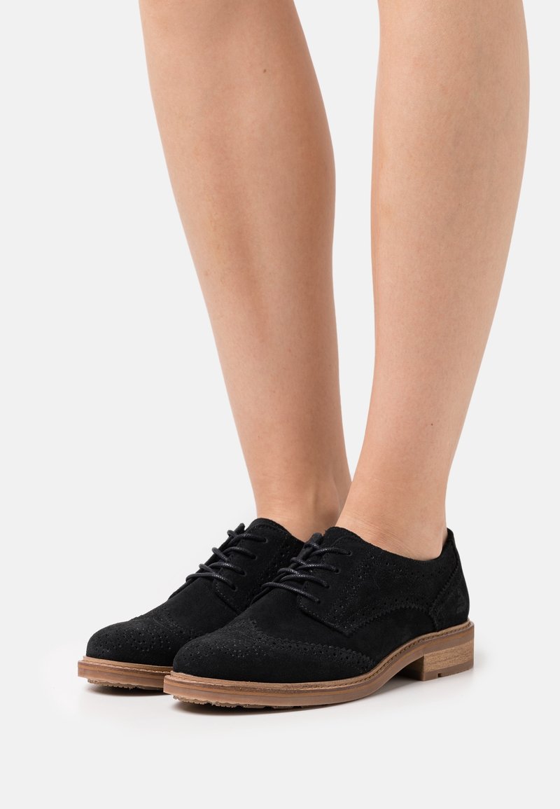 Chaussures brogue en daim noir à lacets avec semelle en bois portées sur jambes nues sur fond blanc.