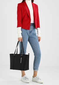 Černá taška typu tote s dlouhými uchy, hladkou texturou, s bílým logem "VALENTINO", doplněná červeným sakem, rolákem a modrými džínami.
