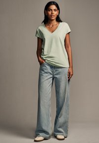 Hellgrünes V-Ausschnitt-T-Shirt aus weichem Stoff, kombiniert mit lockeren, hochtaillierten hellblauen Jeans. Schuhe in neutralen Farben runden das Outfit ab.