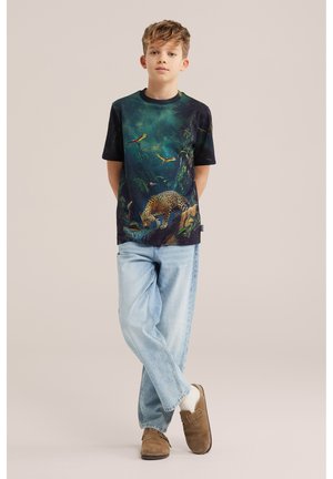 Dječak stoji sa prekriženim nogama noseći tamnu majicu s temom džungle na kojoj su leopard i ptice, svijetloplave traperice, bijele čarape i smeđe cipele.