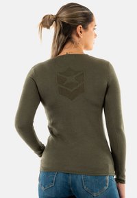 Chemise à manches longues olive, présentant un motif géométrique texturé dans le dos, au design ajusté et avec des poignets côtelés. Associée à un jeans bleu.