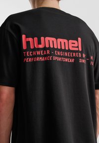 Svart t-shirt med kort ärm, med en djärv röd grafisk tryck på baksidan som inkluderar varumärkesnamn och beskrivning.