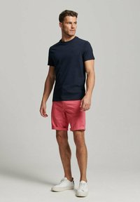 Hombre de pie que lleva una camiseta azul marino, pantalones cortos rosa coral y zapatillas blancas, mirando de lado contra un fondo sencillo.