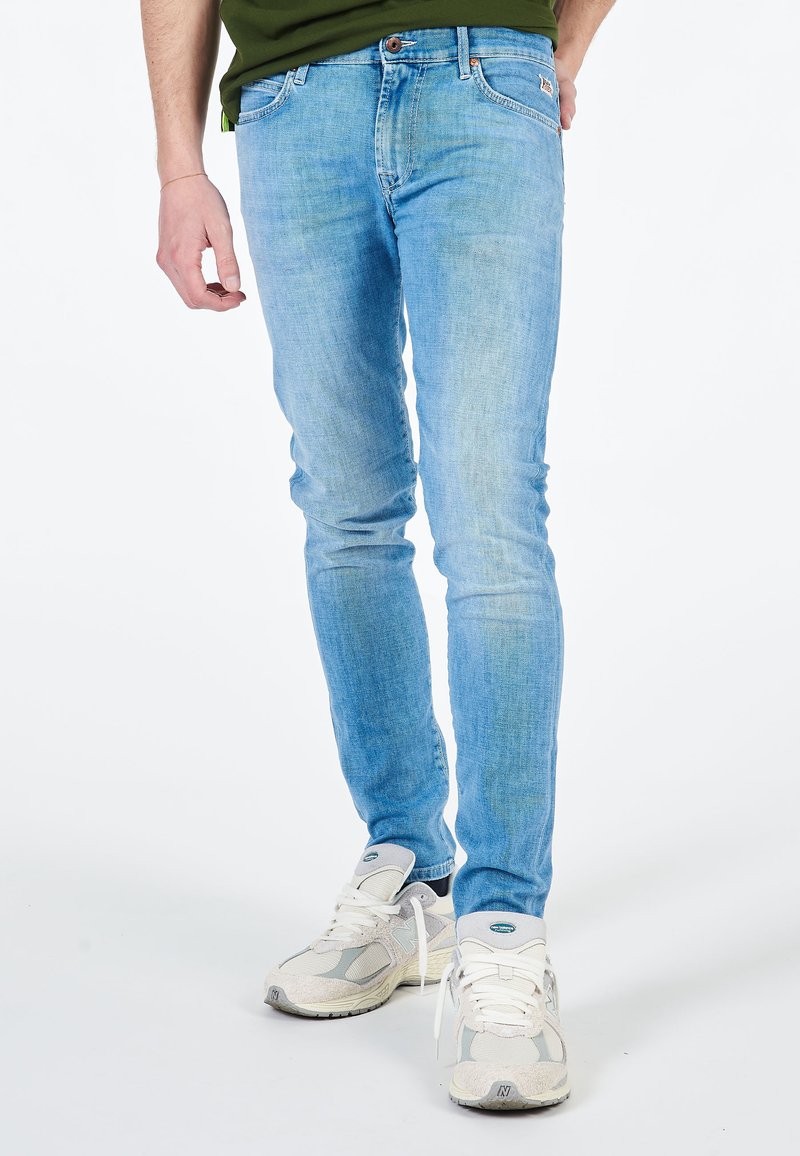 Jeans slim-fit azzurri chiari con una texture sbiadita, cinque tasche e zip, abbinati a scarpe da ginnastica grigie.