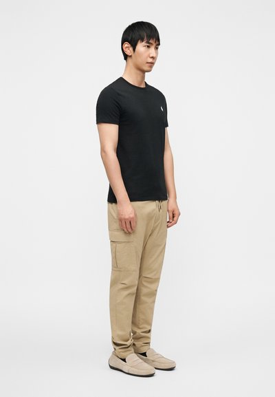 Μαύρο βαμβακερό μπλουζάκι με στρογγυλή λαιμόκοψη, ταμπά παντελόνι εργασίας με πλαϊνές τσέπες και μπεζ παπούτσια τύπου slip-on. Απλός και πρακτικός σχεδιασμός.