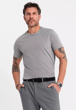 Šedé tričko s kruhovým výstřihem z měkké tkaniny, přiléhavý střih, nošené s šedými kalhotami a černým páskem. Viditelné tetování na levém rameni.