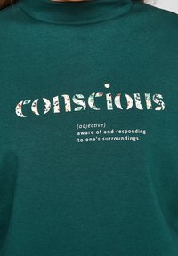 Tiefblaues Rollkragenshirt mit dem Wort "bewusst" in floral gemusterten Buchstaben, darunter in weißen Schrift die Definitionen auf glattem Stoff.