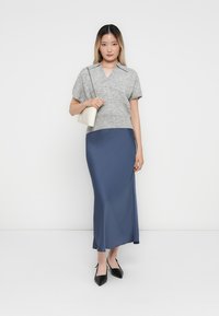 Top in maglia grigia a maniche corte abbinato a una gonna lunga in satin blu. La modella tiene in mano una piccola borsa bianca; i tacchi a punta neri completano l'outfit.